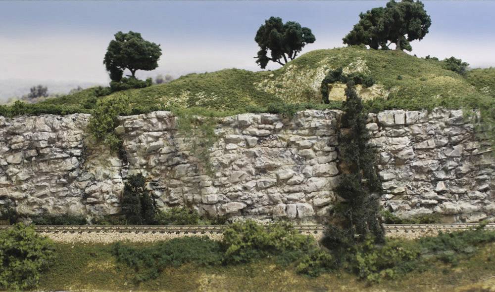 Landscape with a rocky cliff, grass-covered hills and trees; a railway line runs along the cliff in the foreground.