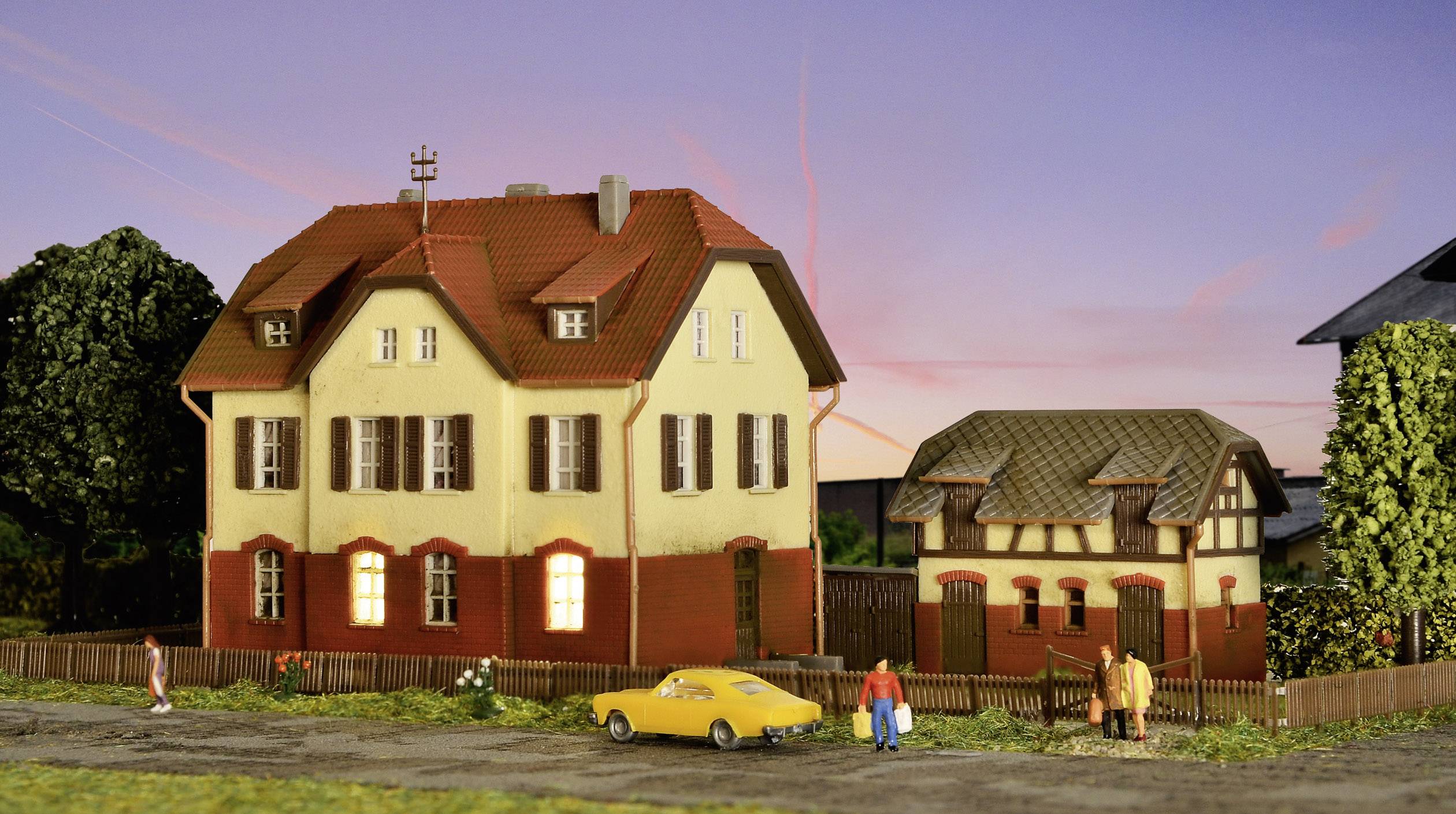 A large model house with a red roof stands next to a smaller building. People and a yellow car are positioned in front of it.