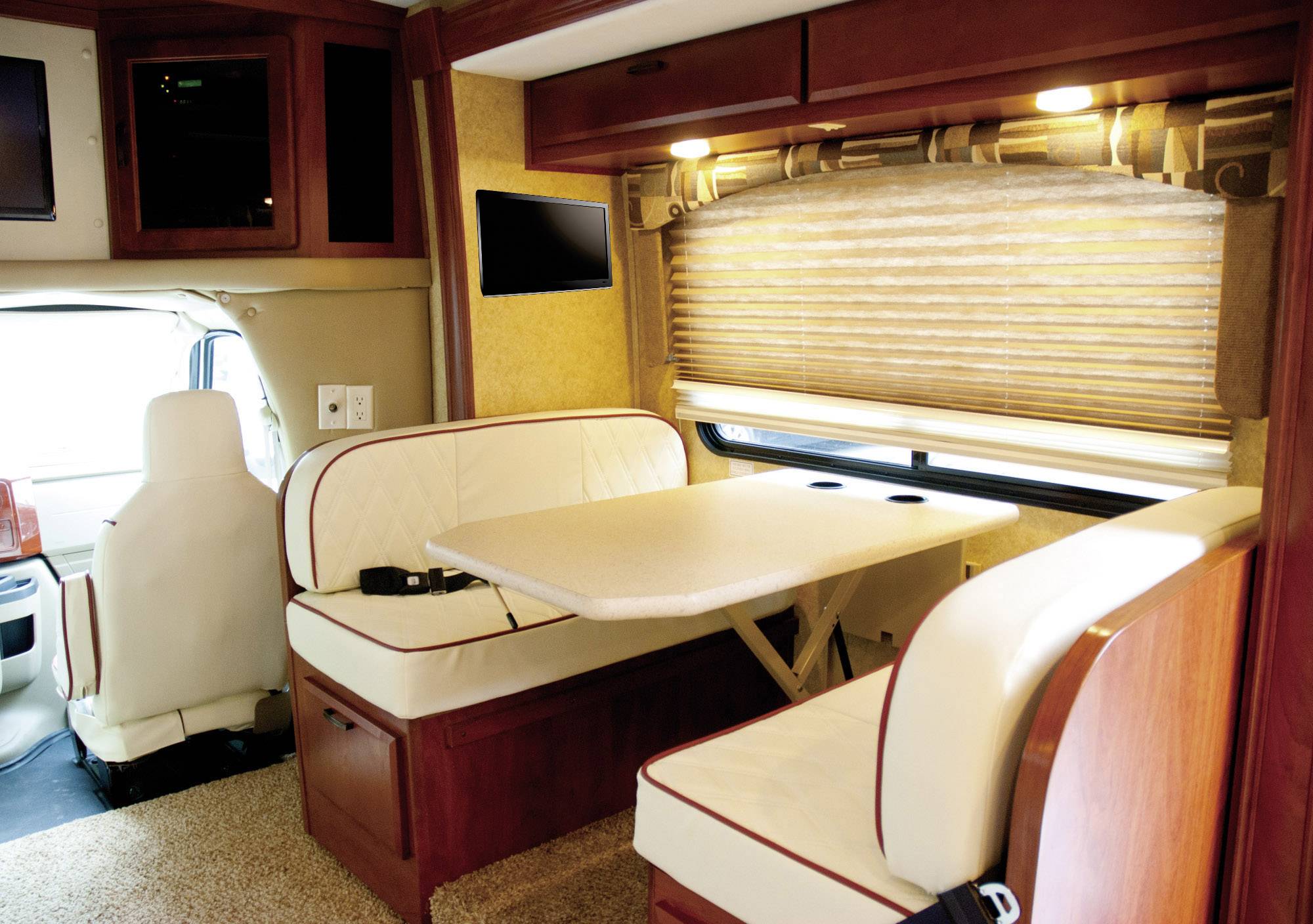A motorhome interior with a dining table between two bench seats. Daylight streams in through a window with wooden venetian blinds.