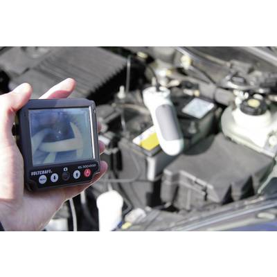 A person is holding a diagnostic device with a screen, displaying the internal condition of a car part. The engine compartment is visible in the background.