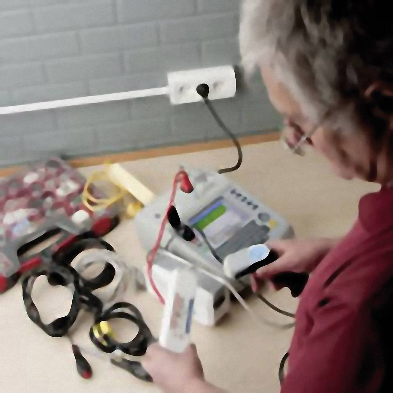 A person is measuring blood pressure using a blood pressure monitor and cables in a room. In the background, toolboxes and plug sockets are visible.