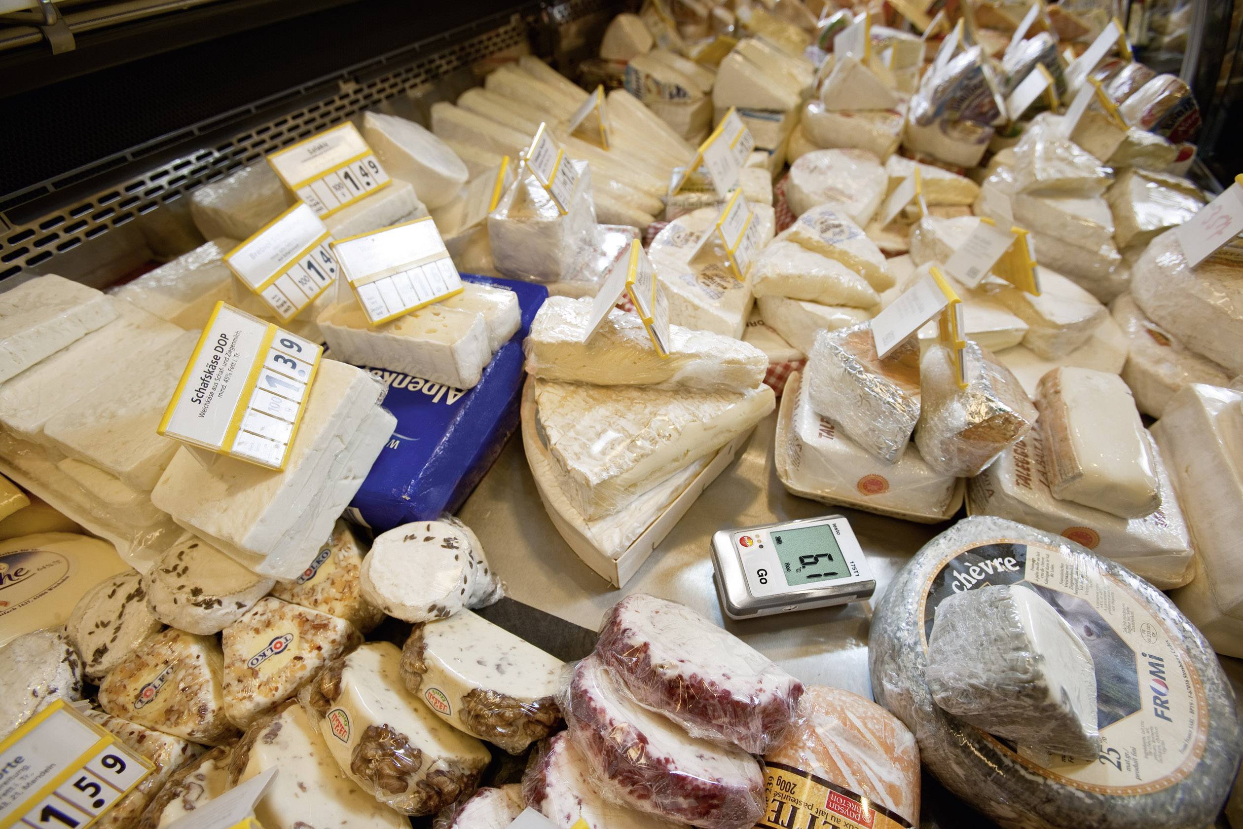 Various types of cheese are displayed on a counter in a supermarket, with price and weight labels on each individual piece.