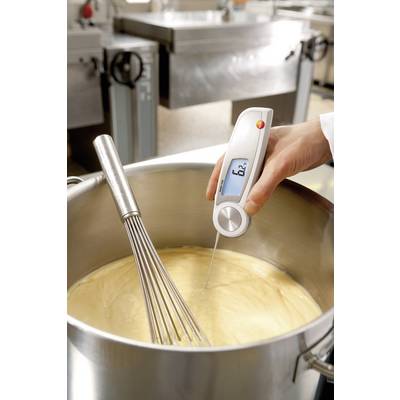 A hand is holding a thermometer measuring the temperature of a yellow sauce in a saucepan next to a whisk.