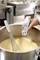 A hand is holding a thermometer measuring the temperature of a yellow sauce in a saucepan next to a whisk.