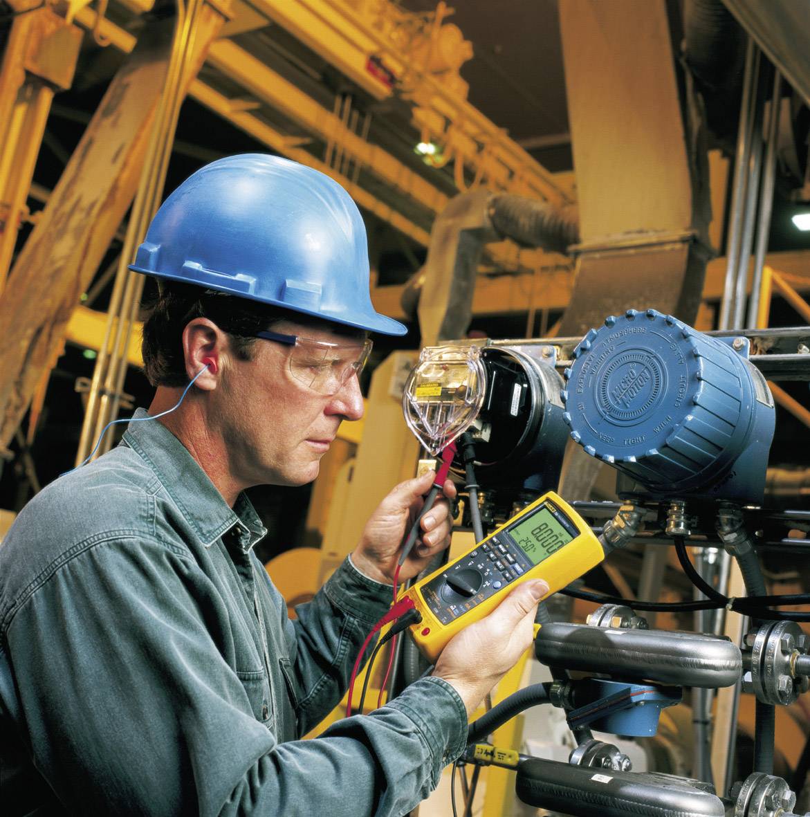 A worker wearing a hard hat and safety glasses is measuring the pressure on an industrial device using a digital measuring instrument.