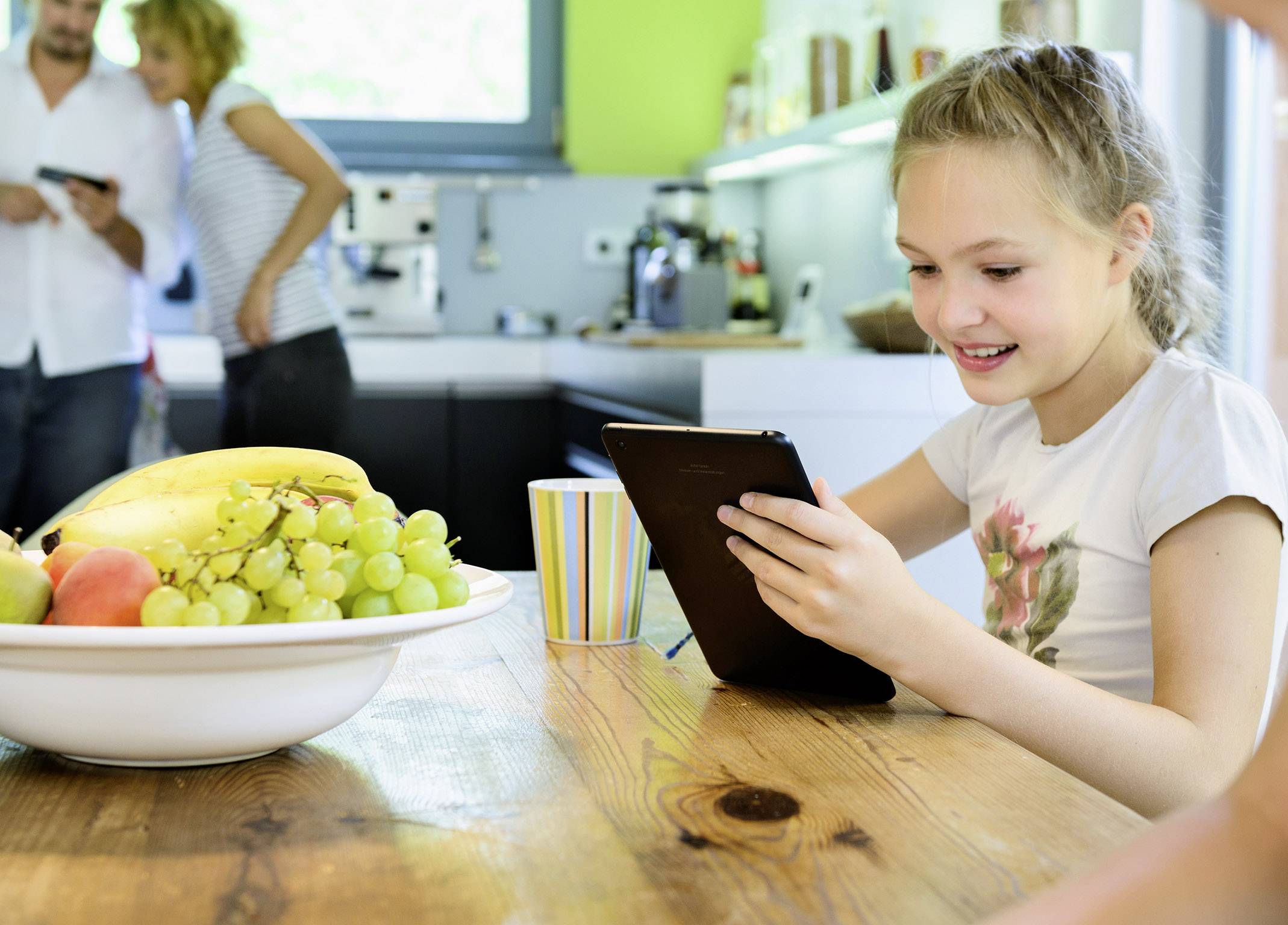 A girl is sitting at the table using a tablet, with a couple visible in a modern kitchen in the background. A plate of fruit is on the table.