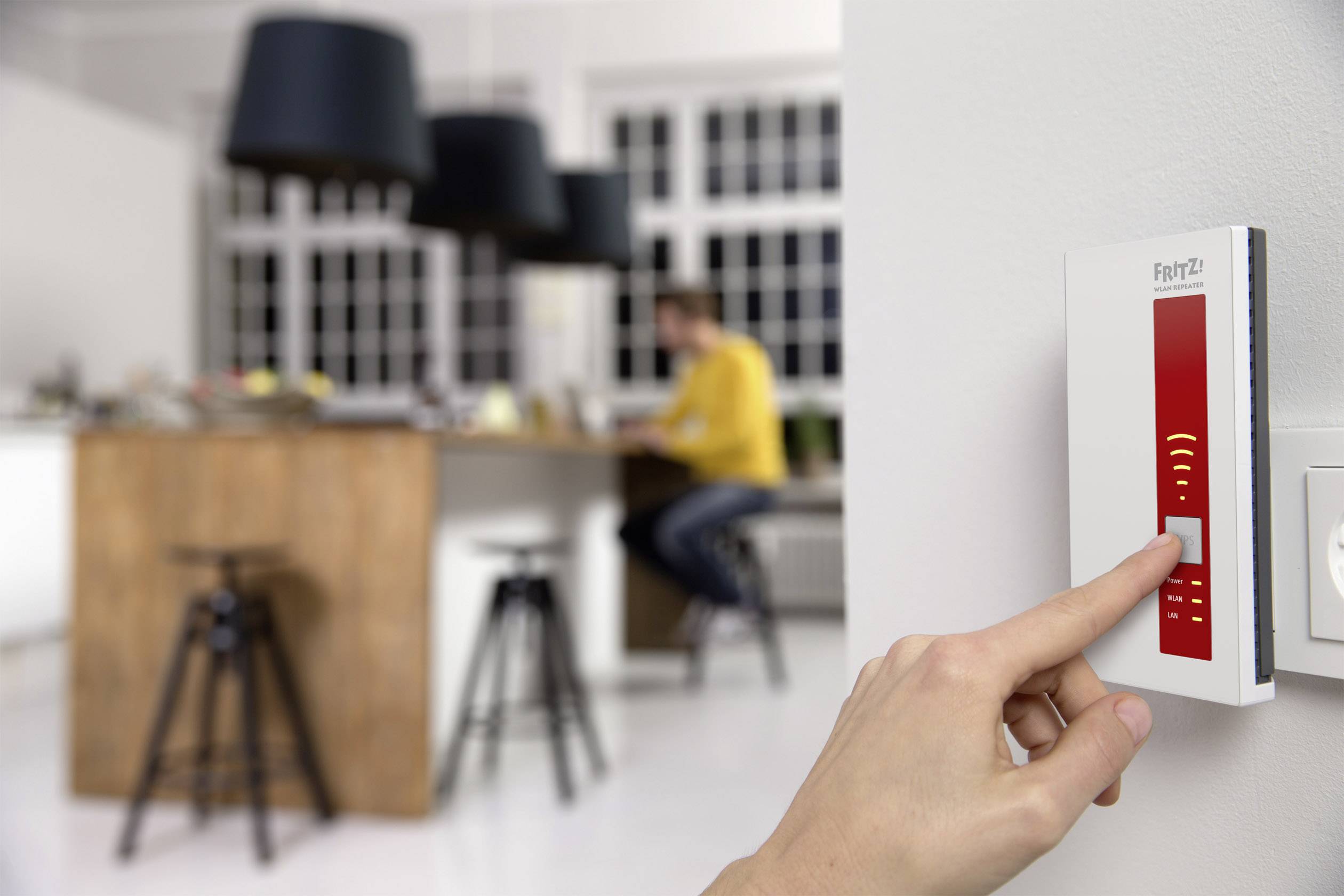 A hand is operating a white and red smart home device on a wall. In the blurred background, a person can be seen in a modern kitchen.