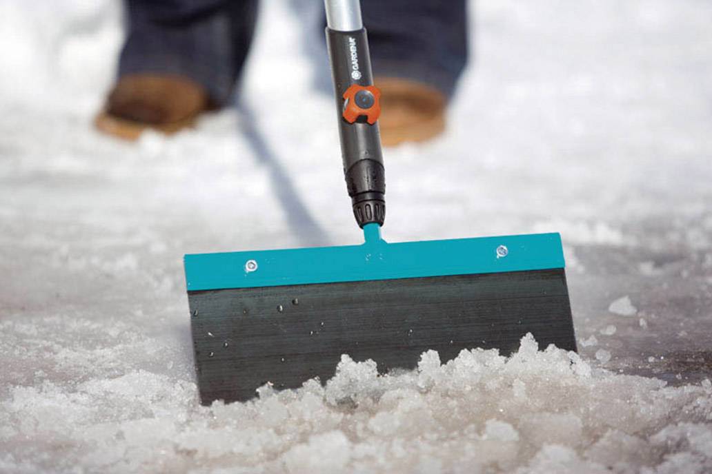 A person is clearing snow from a pavement using a snow shovel. The blue shovel blade is visible in the foreground.