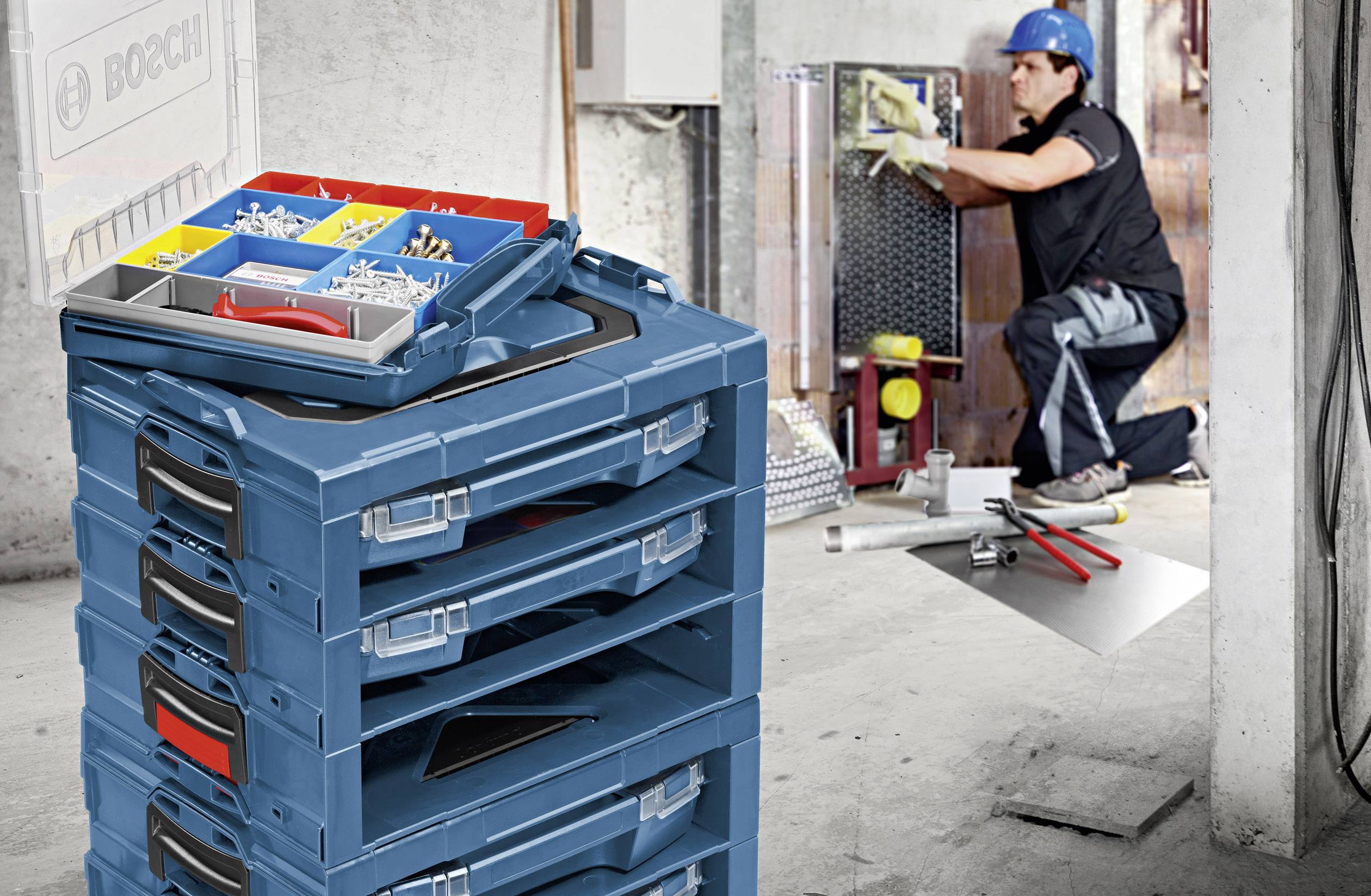 A tradesman is installing devices on a wall. In the foreground, there is a stack of blue toolboxes with sorted small parts.