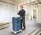 A worker in protective clothing and a red hard hat is pulling a trolley with stacked crates in a timber-frame building.