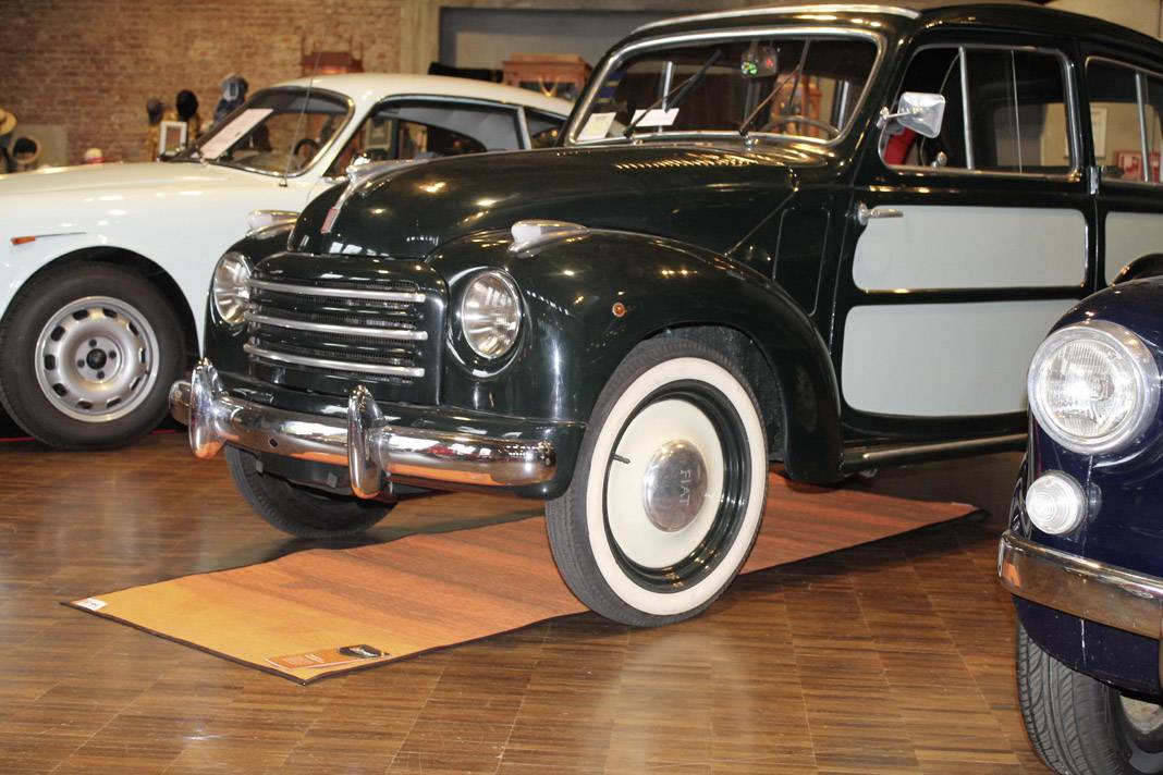 An antique, dark green car is displayed in a hall on a wooden floor. Other classic cars are visible beside the vehicle.