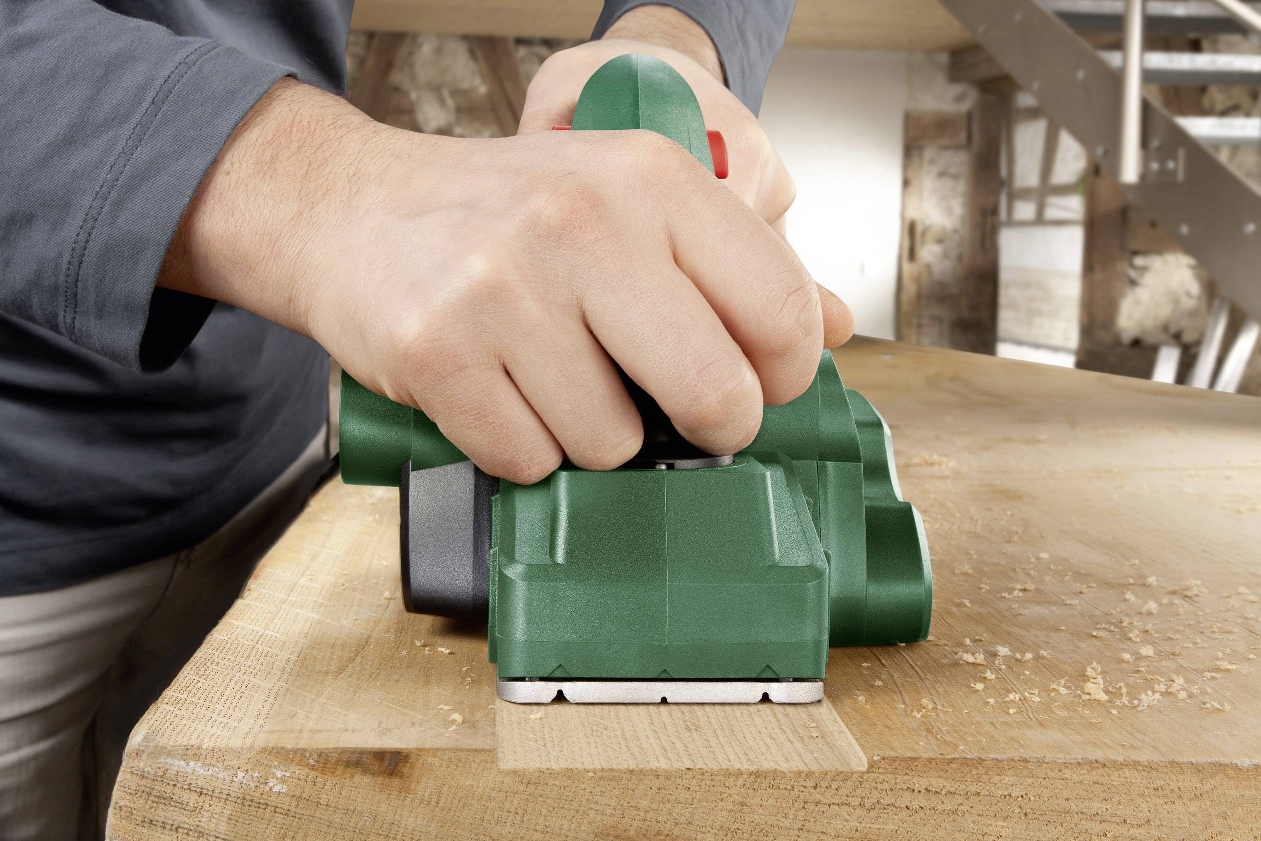 A person is using a green electric plane to smooth the surface of a wooden board in a workshop.