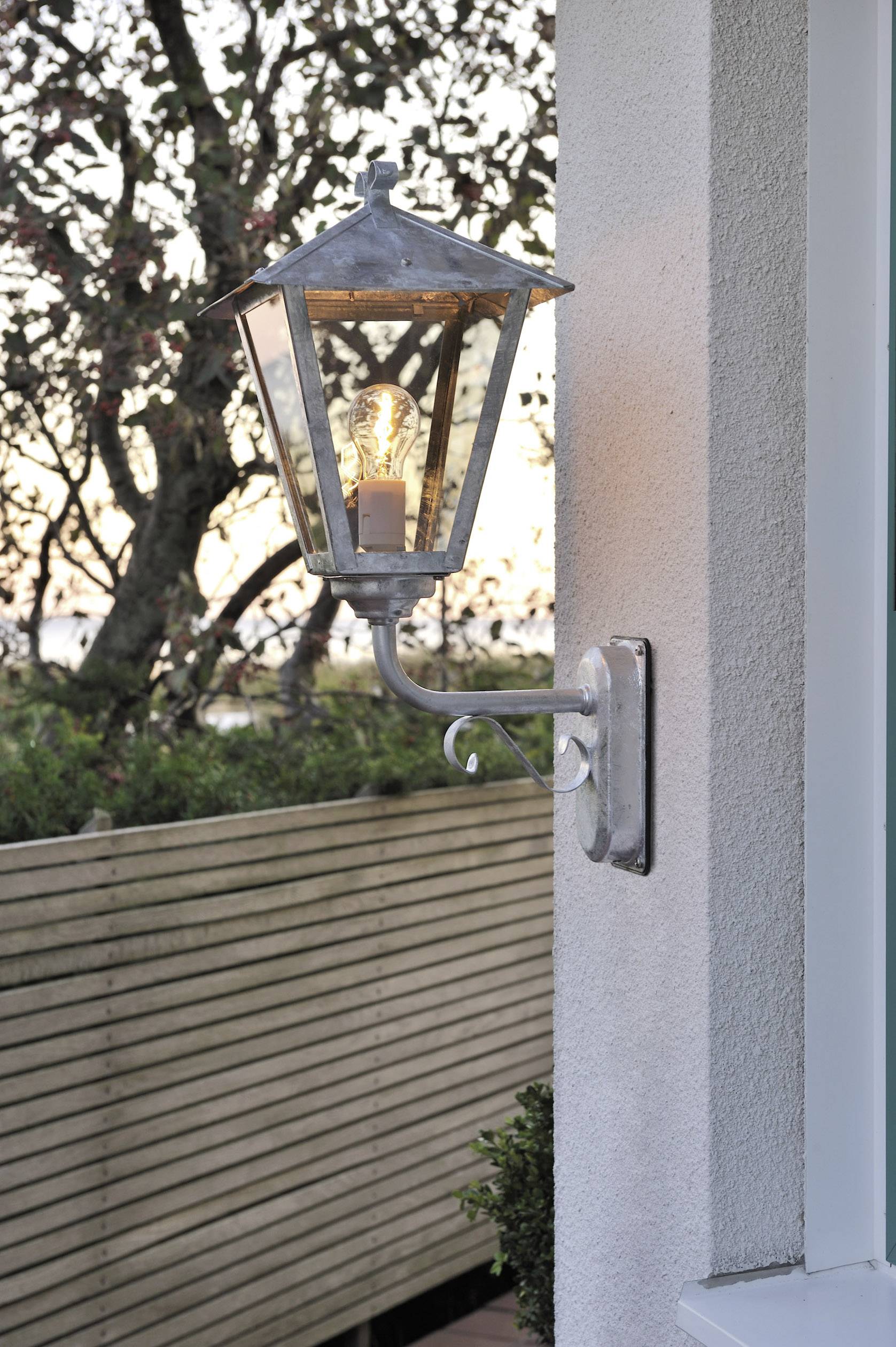 Wall light in a traditional style illuminates at dusk. It is mounted on a house wall, with a tree in the background.