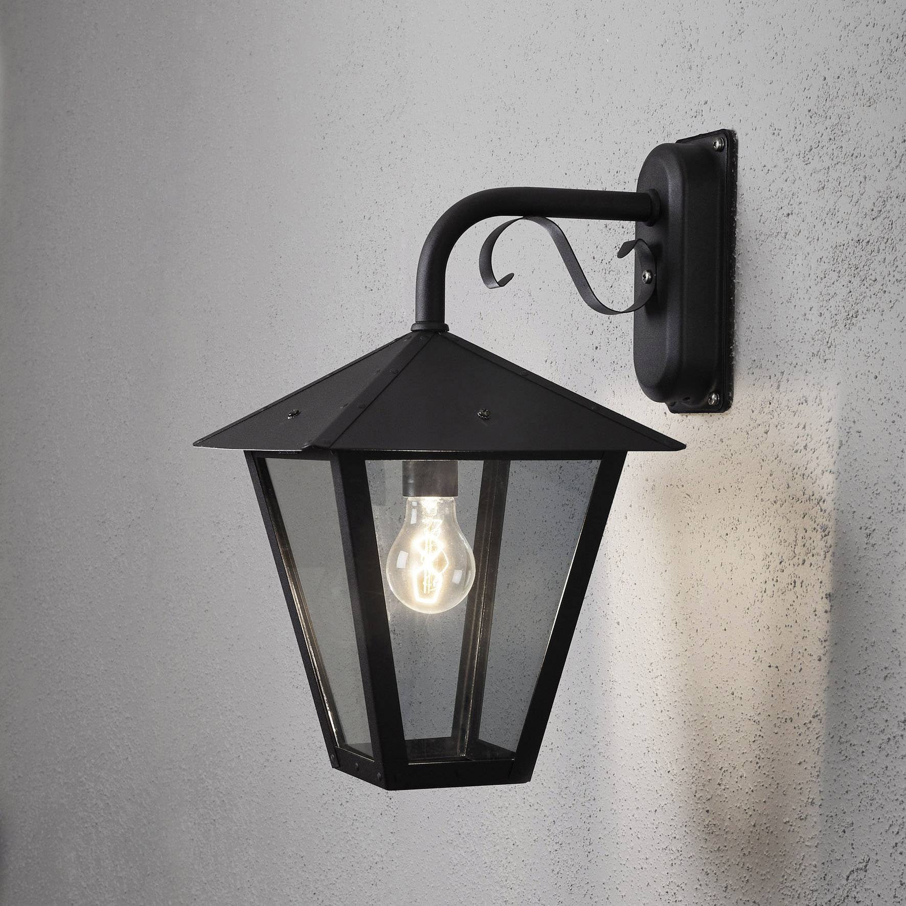 Black wall light with a pointed hood, mounted on a grey wall, illuminated by a clear light bulb inside.