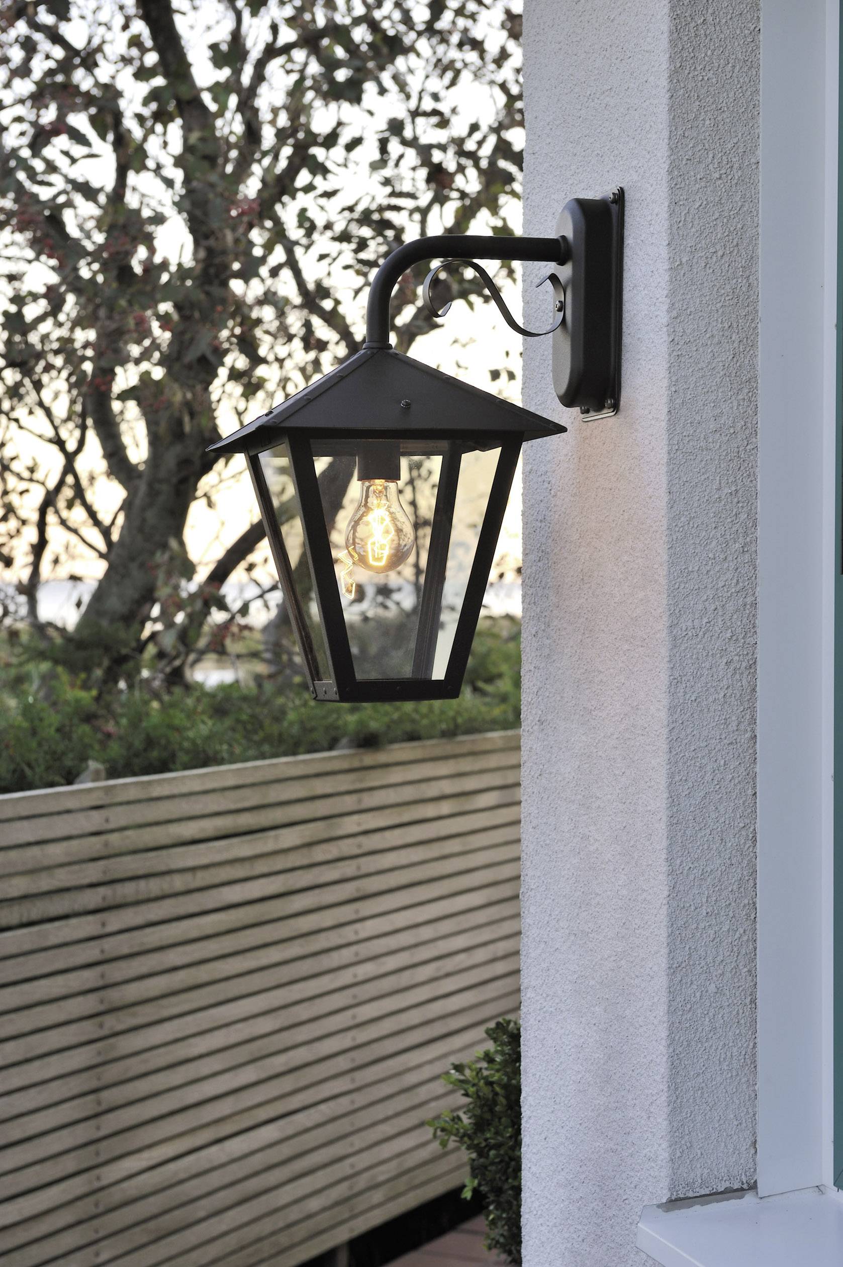 Exterior light on a house wall, switched on at dusk. In the background, a wooden fence and trees are visible.