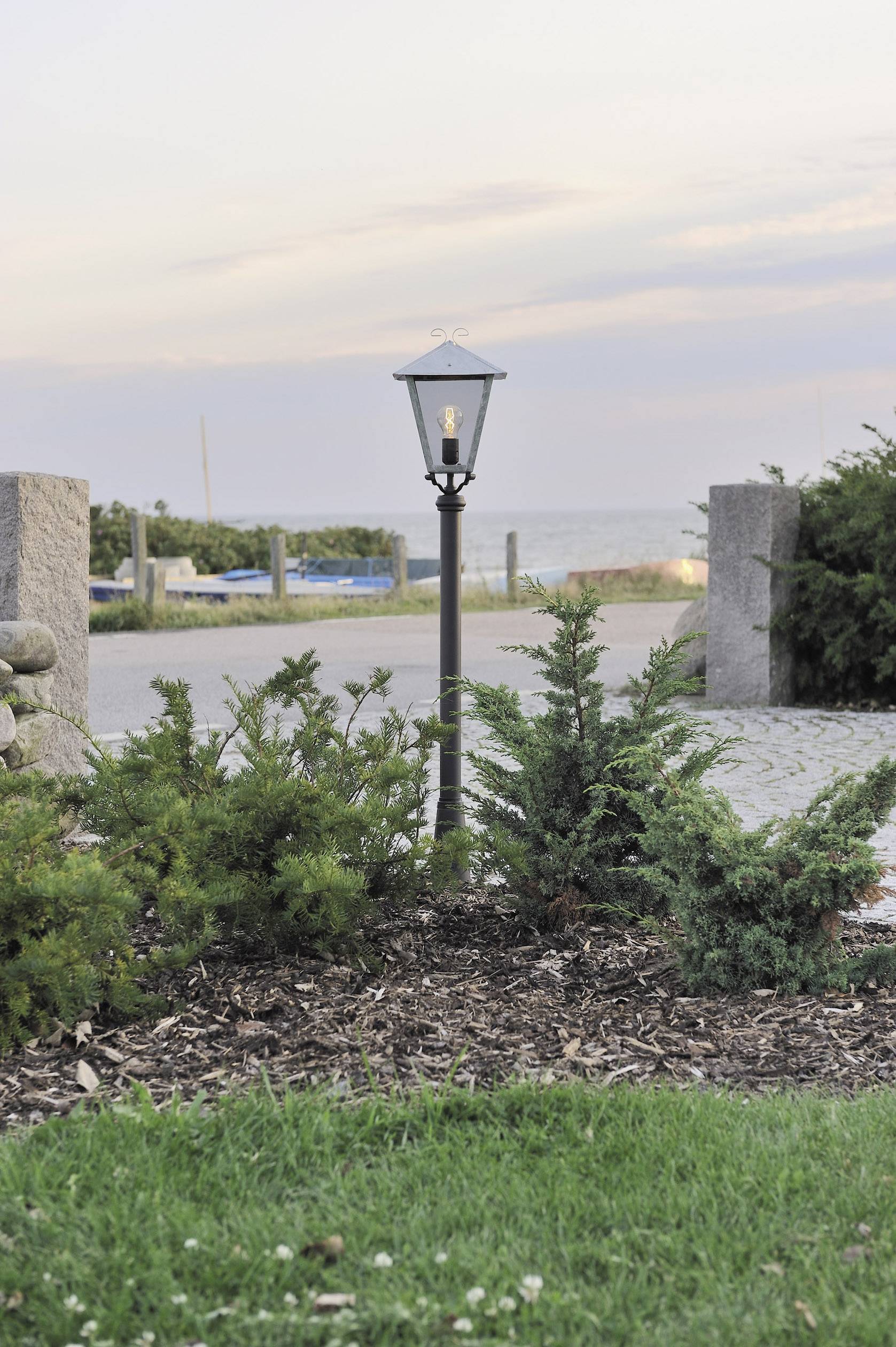 A garden lantern illuminates a path near a coastline. Small shrubs are in the foreground, with the sea visible in the background.