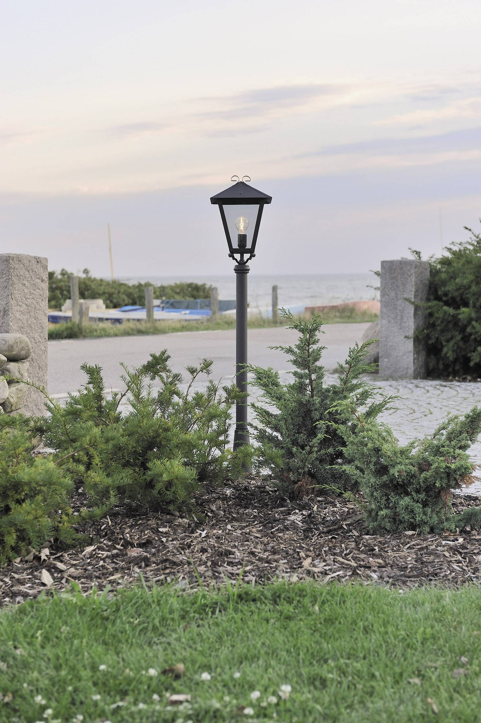 A lantern stands in a garden bed, surrounded by small shrubs, with a paved pathway in the background and a view of the sea.