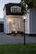 A lit path leads to a two-storey wooden house. Two lanterns stand alongside the gravel path, and the windows are softly illuminated.