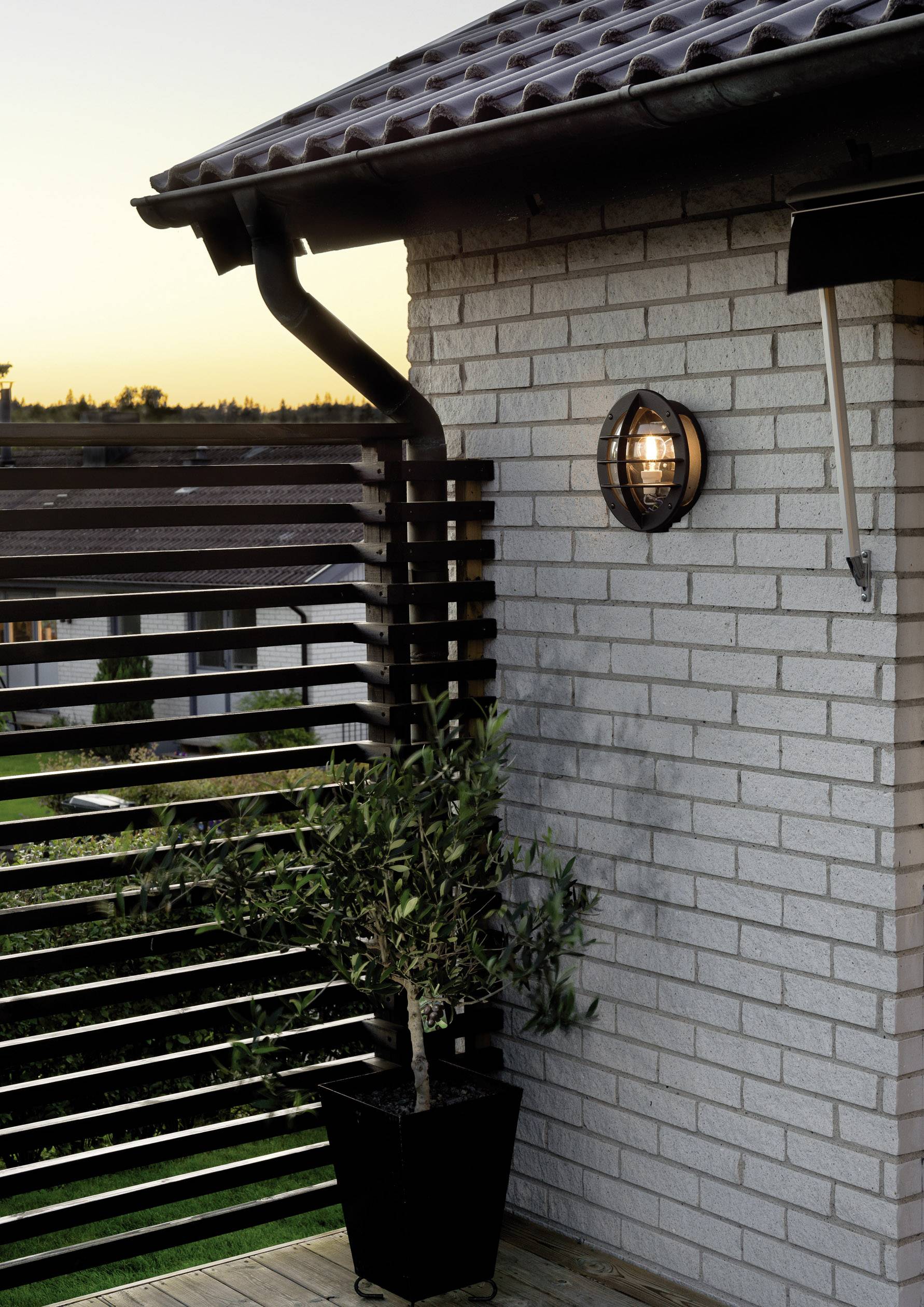 Balcony at sunset with plants, a modern railing and a wall-mounted lantern on a brick house, radiating warm light.