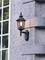 Exterior wall lantern made of black metal with an illuminated light bulb mounted on a textured stone wall beside a window.