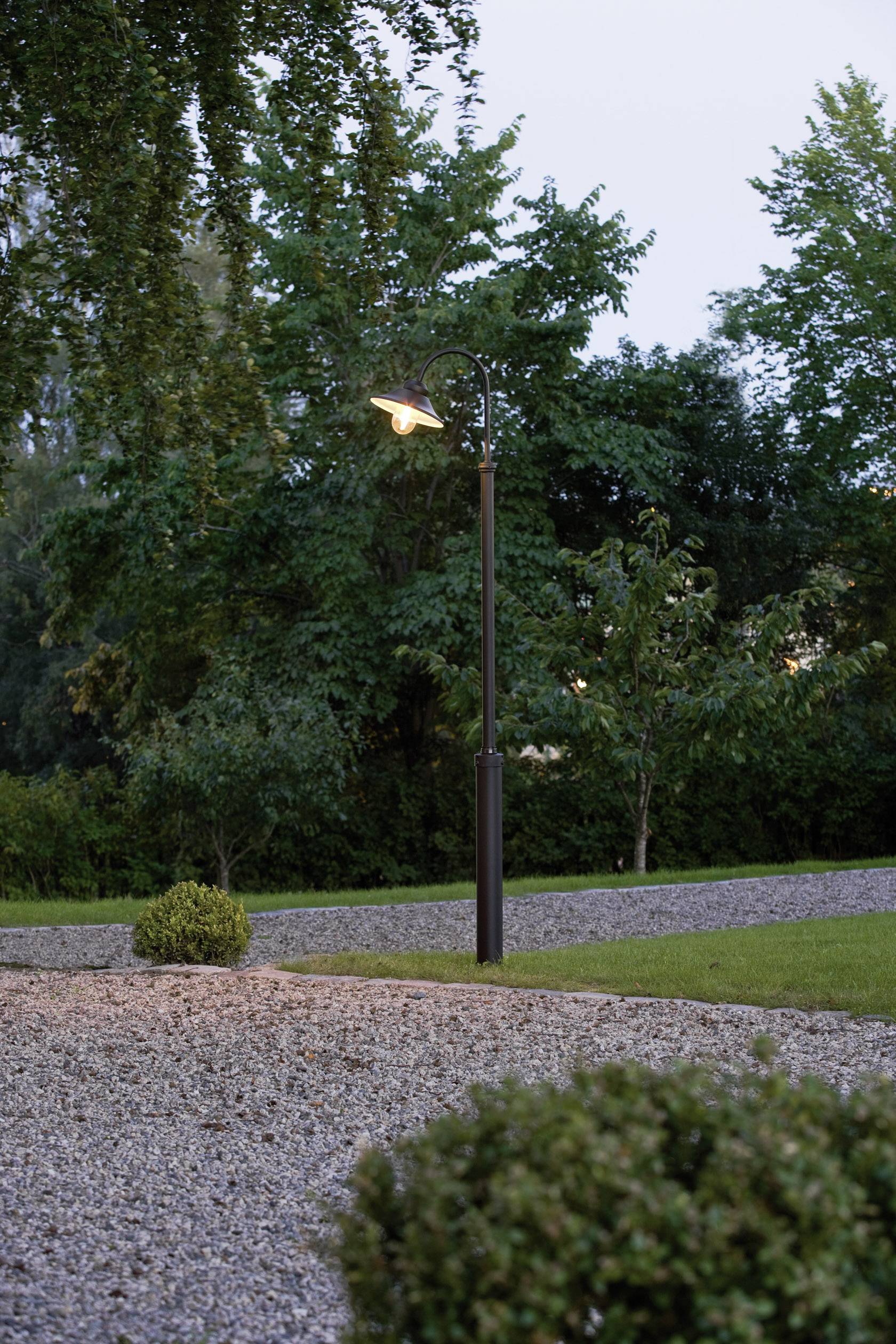 A lit street lamp post stands in a gravel garden, surrounded by green bushes and trees in the background at dusk.