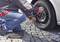 A person is changing a car tyre on a paved path, using a spanner and wearing casual clothing.