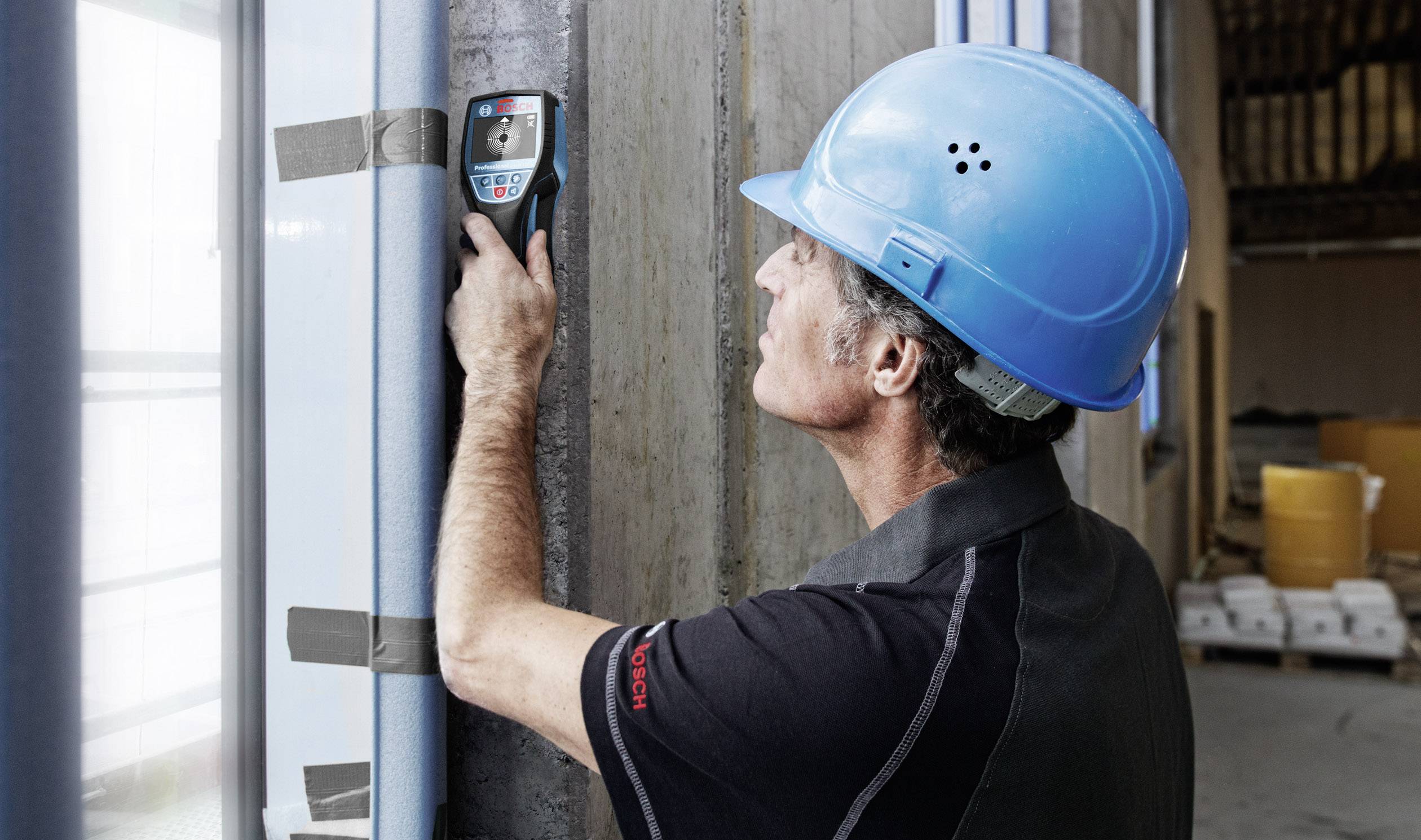 A construction worker is measuring the thickness of a wall with a device. He is wearing a blue hard hat and is focused on taking the measurement.
