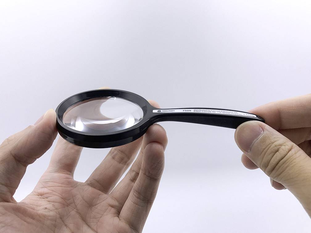 A hand holds a black magnifying glass over an open palm, magnifying details. On a white background without additional objects.