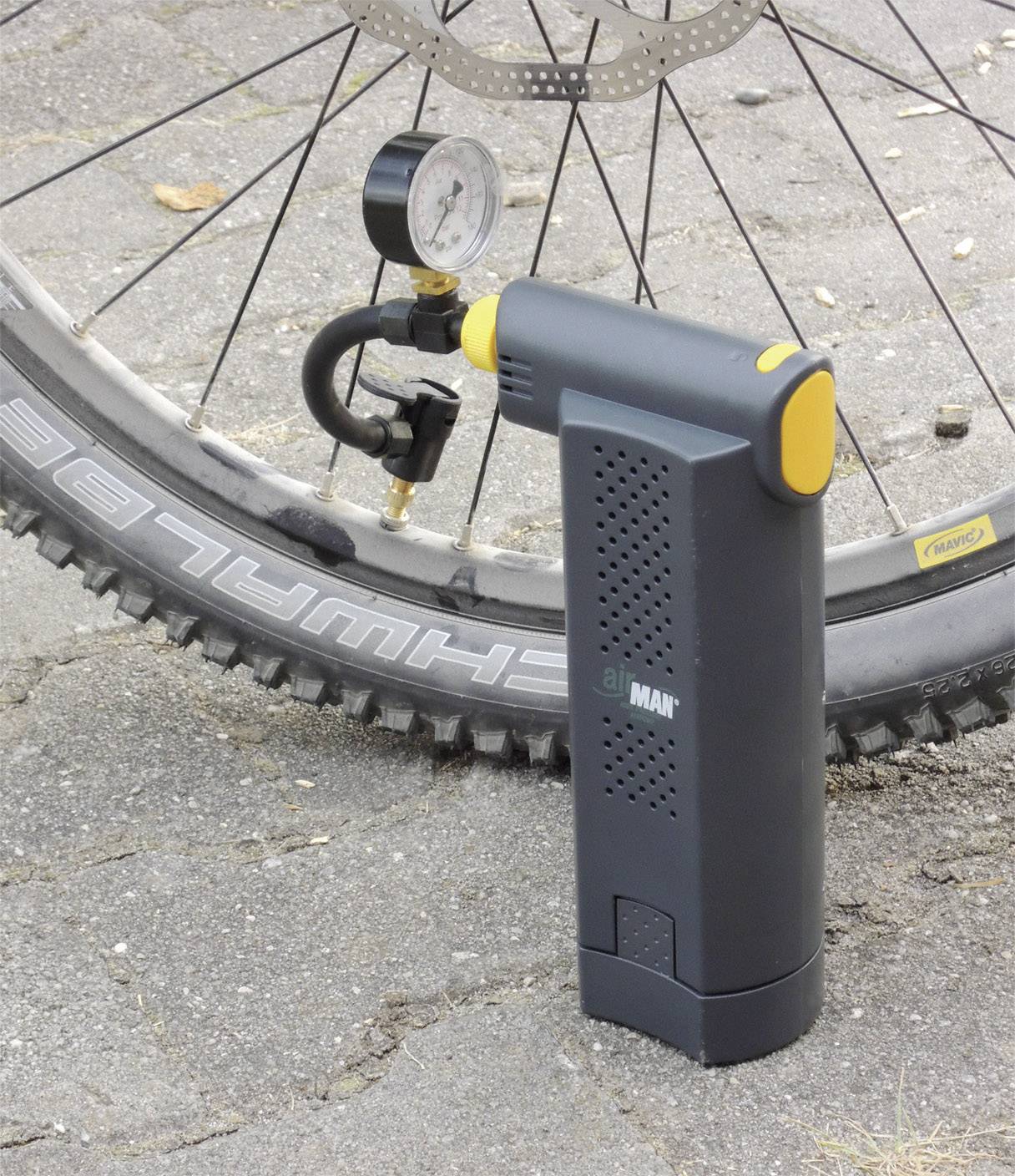 Electric bicycle pump stands next to a bicycle tyre on tarmac. Pump display shows pressure. Tyre has a coarse tread pattern.