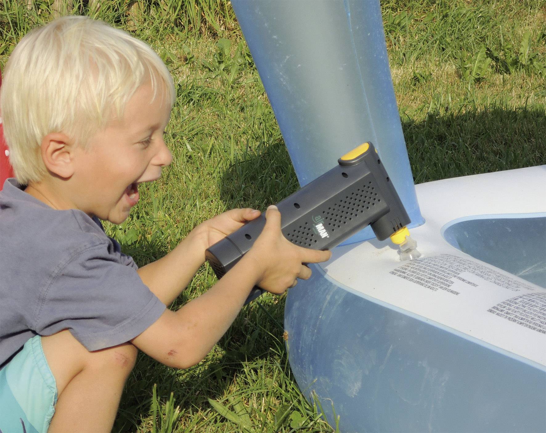 A child sits on grass and pumps air into an inflatable paddling pool using an electric pump. They look joyful and focused.