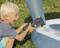 A child sits on grass and pumps air into an inflatable paddling pool using an electric pump. They look joyful and focused.