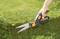 A hand uses garden shears to cut a lawn surface precisely. The environment is green and well-maintained.