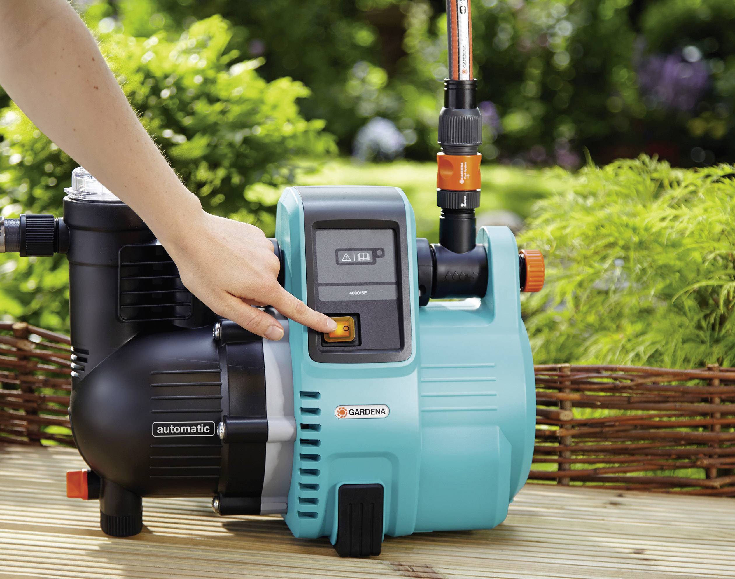 A hand is pressing the button of a blue garden pump, which stands on a wooden surface, surrounded by green plants.