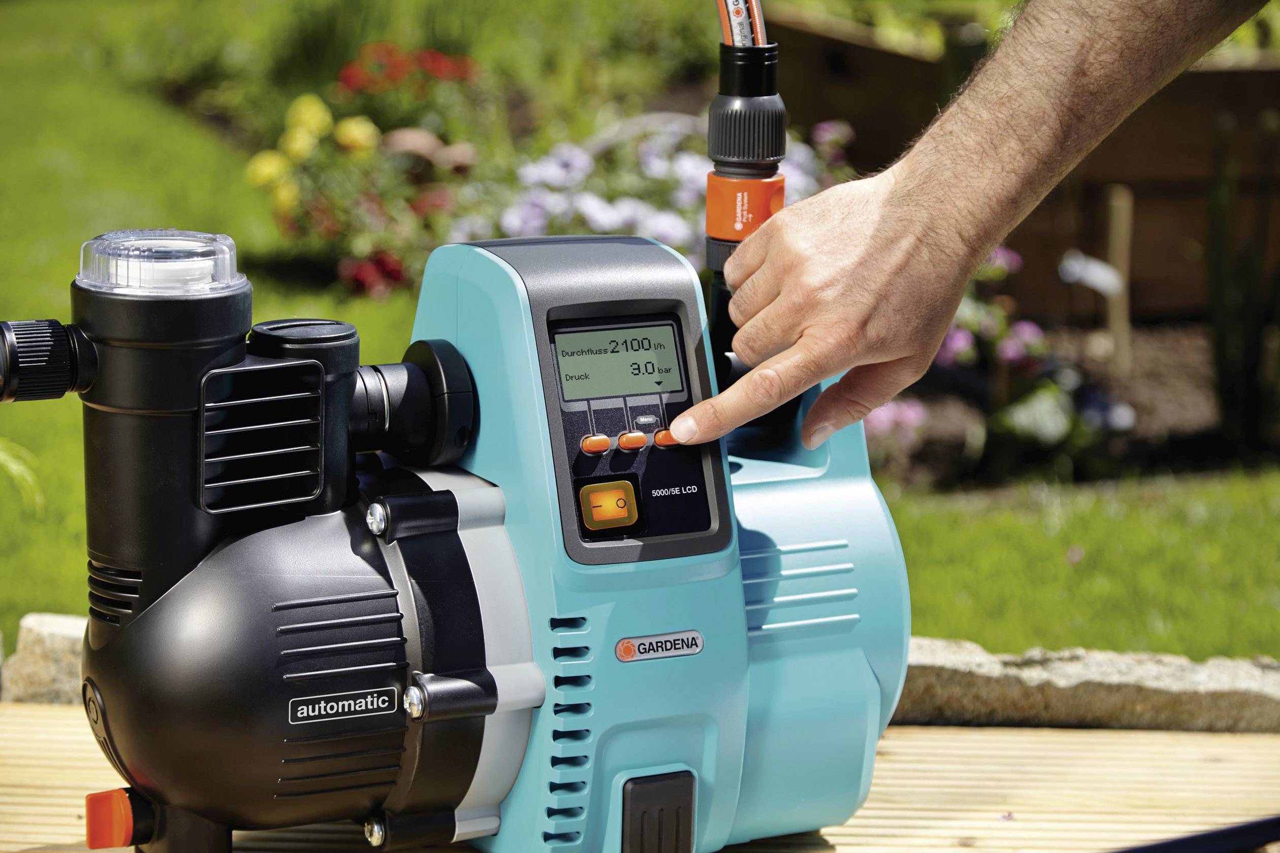 A person is operating a garden pump with a digital display in the foreground. In the background, a blooming garden is bathed in sunlight.