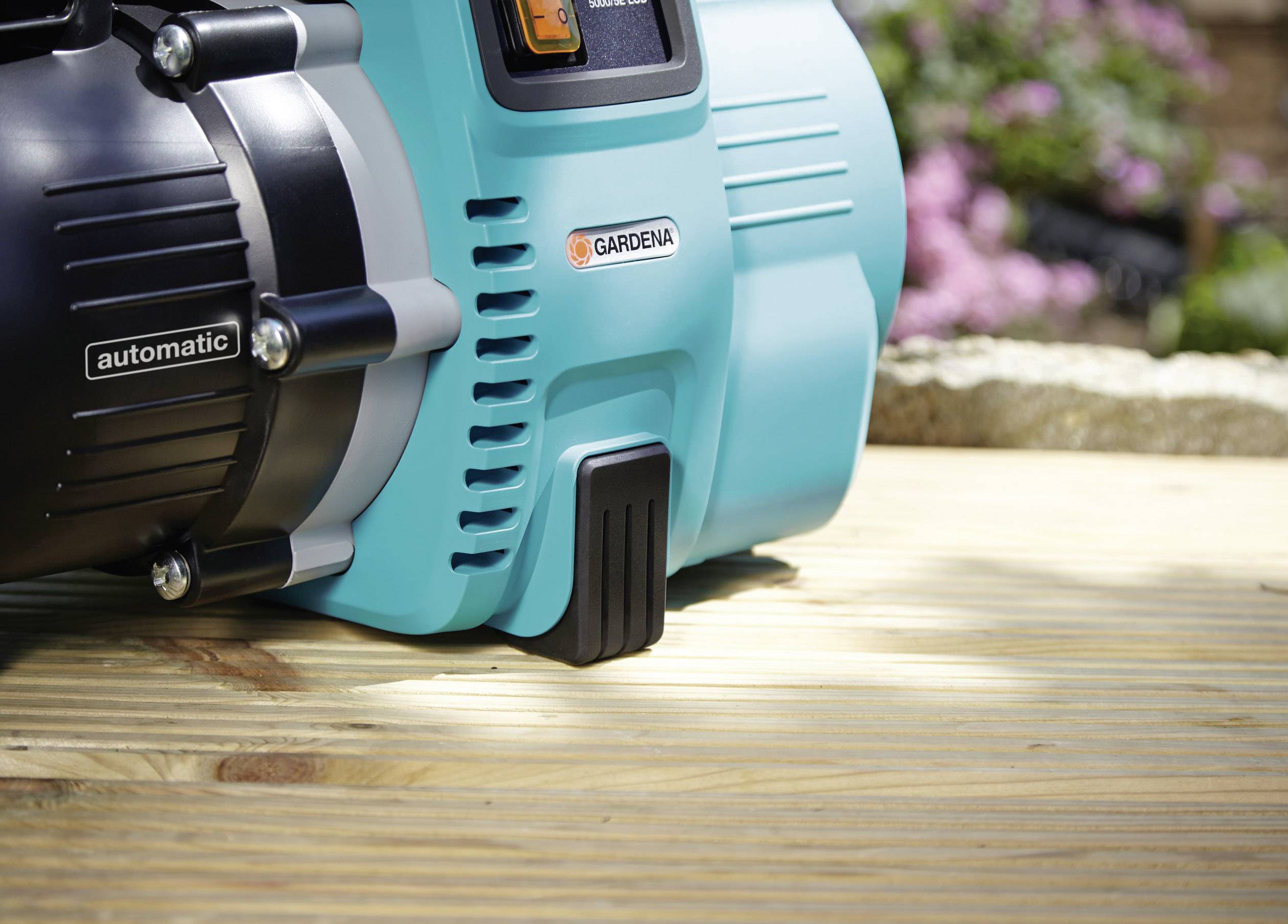 Garden pump by Gardena on a wooden decking, with blurred flowers in the background.