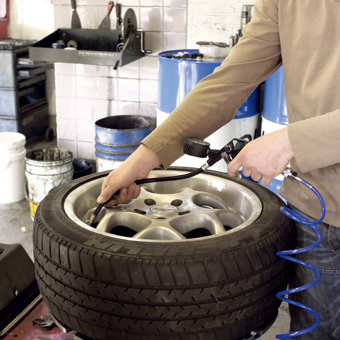 A person is mounting a car tyre in a workshop, holding a tool in their hand. Tools and tyres are visible in the surrounding area.