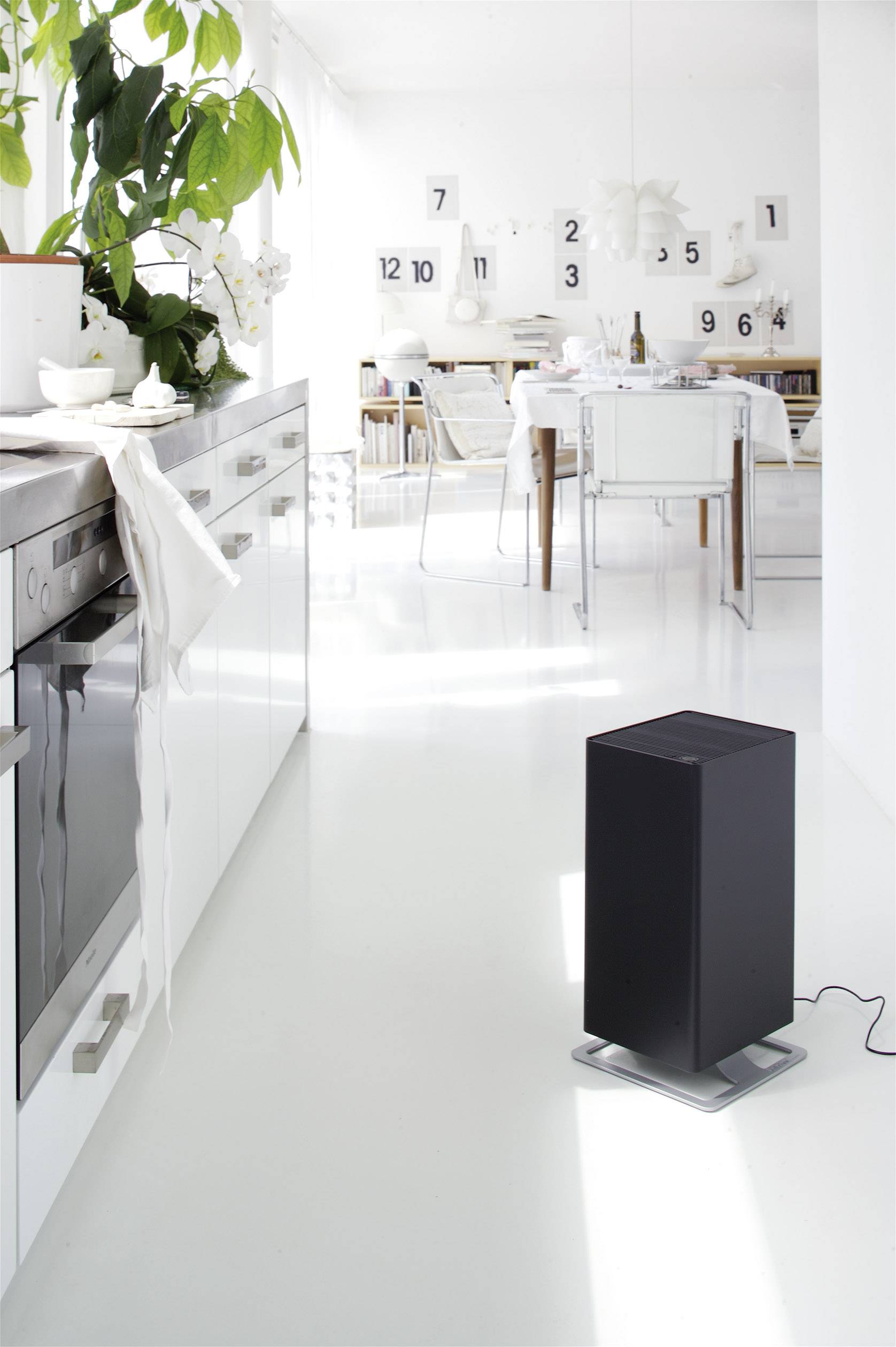 Black air purifier stands on white flooring in a bright, modern kitchen. In the background, a dining area with white furniture.