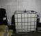 A large white plastic tank in a metal frame stands in a storage room. To the left are cardboard boxes and a black cylinder.