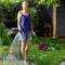 A woman is watering plants with a green hose in a garden. A pump stands on the lawn beside her.