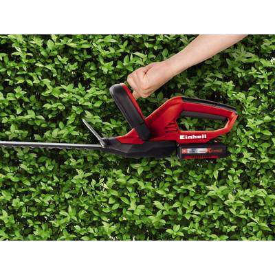 A red hedge trimmer in operation in a hedge. A hand grips the handle firmly. Green leaves are visible in the background.