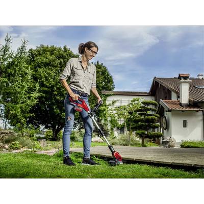 A woman in casual clothing is trimming the lawn beside a house using an electric lawn trimmer. Trees are visible in the background.