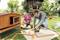 A man is building a rabbit hutch in the garden with a girl. The man is using a tool while the girl is holding a rabbit.