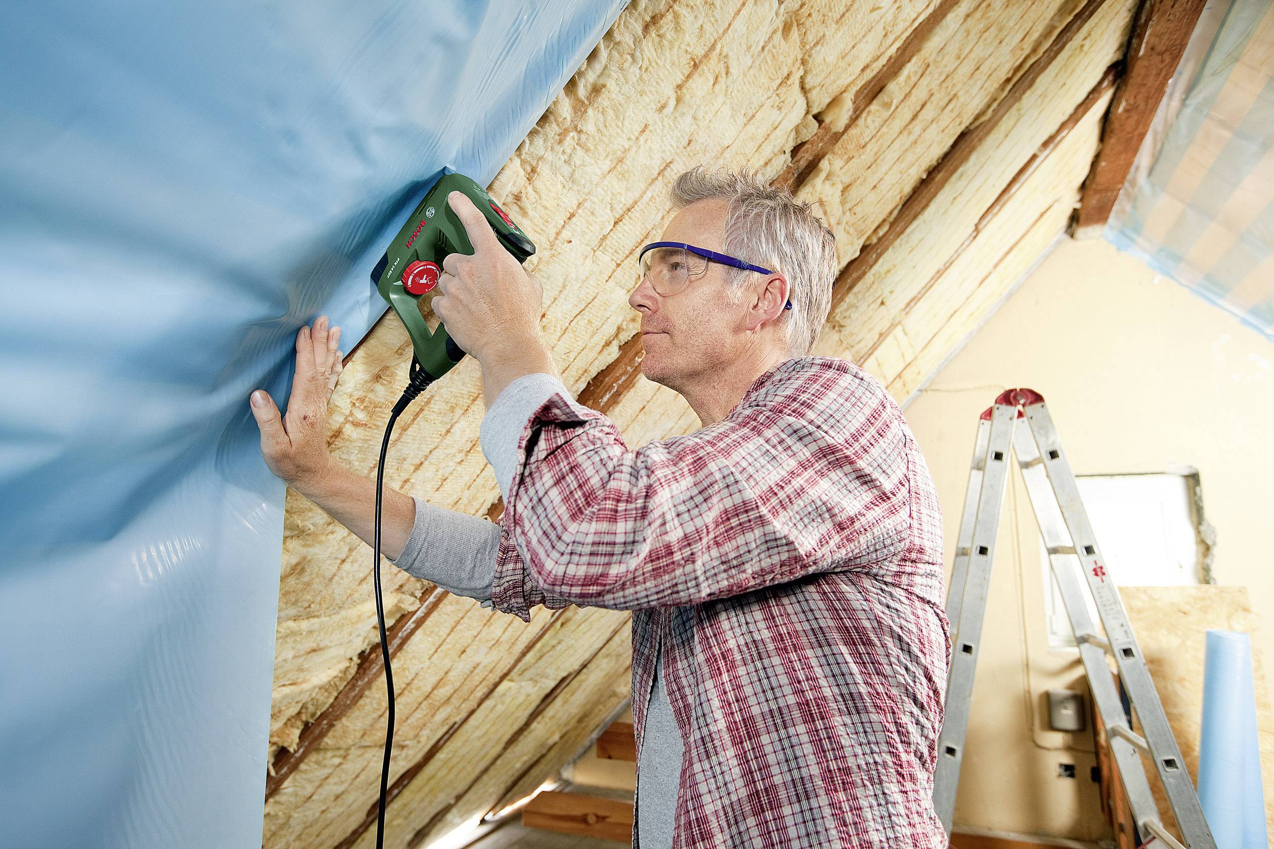 A man is attaching a blue plastic sheet to wooden beams using an electric staple gun in an attic. A ladder is standing in the background.