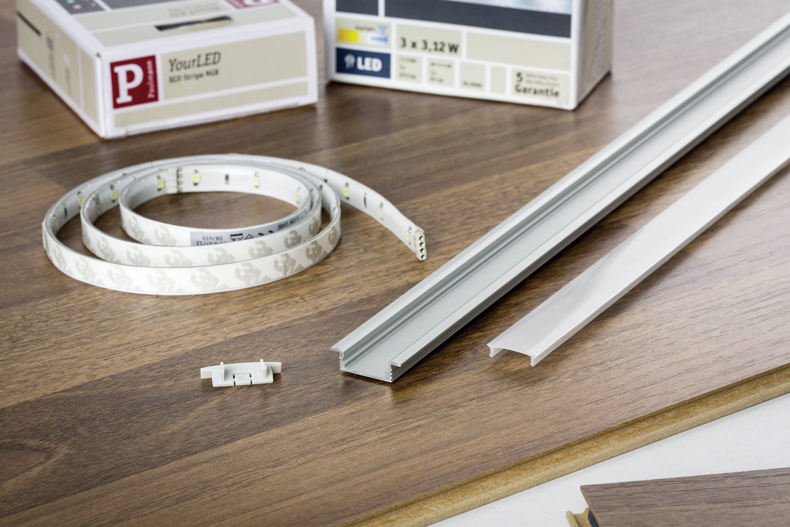 LED lighting set on a wooden table: LED strip, rail, and connecting element. Background: Boxes with product information.