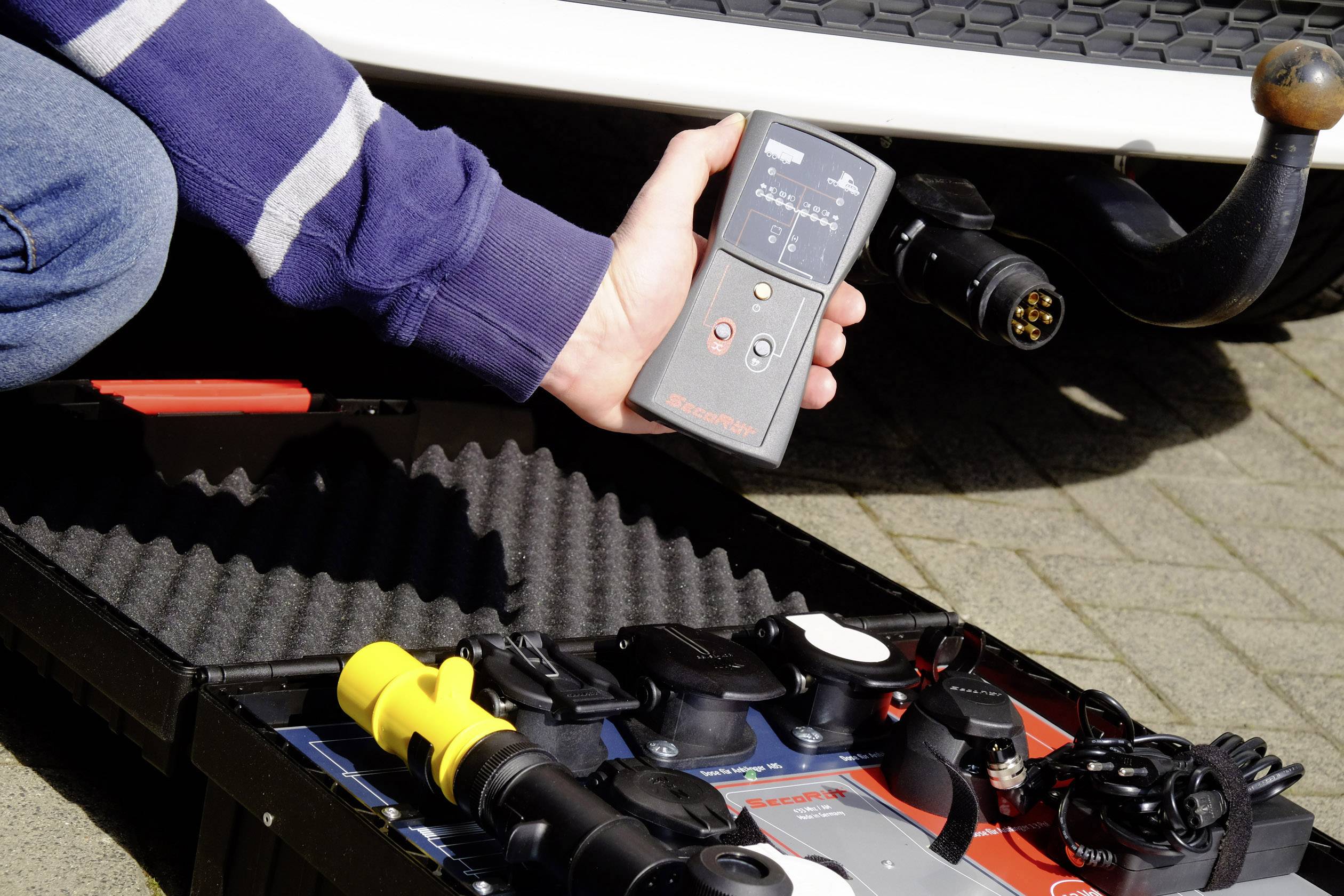 A hand is holding a diagnostic device next to a trailer plug. Beside it lies an open toolbox with various trailer adapters.