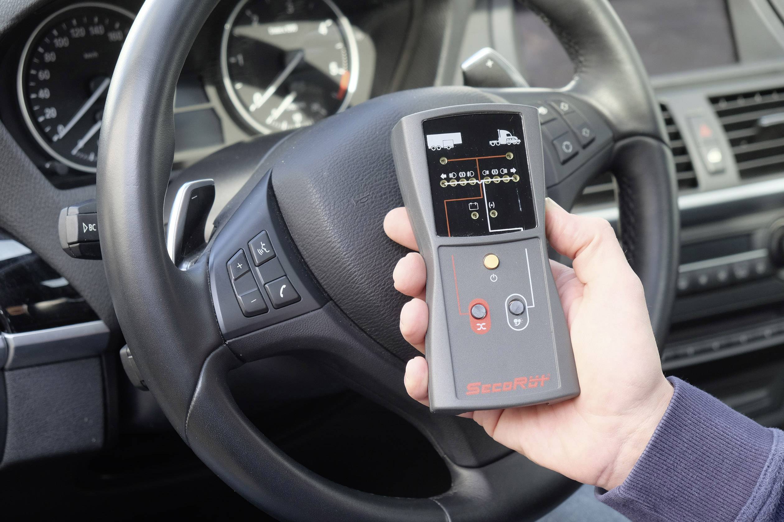 A person is holding an electronic device in a car which could be used for diagnostic or control purposes. A steering wheel is visible in the background.
