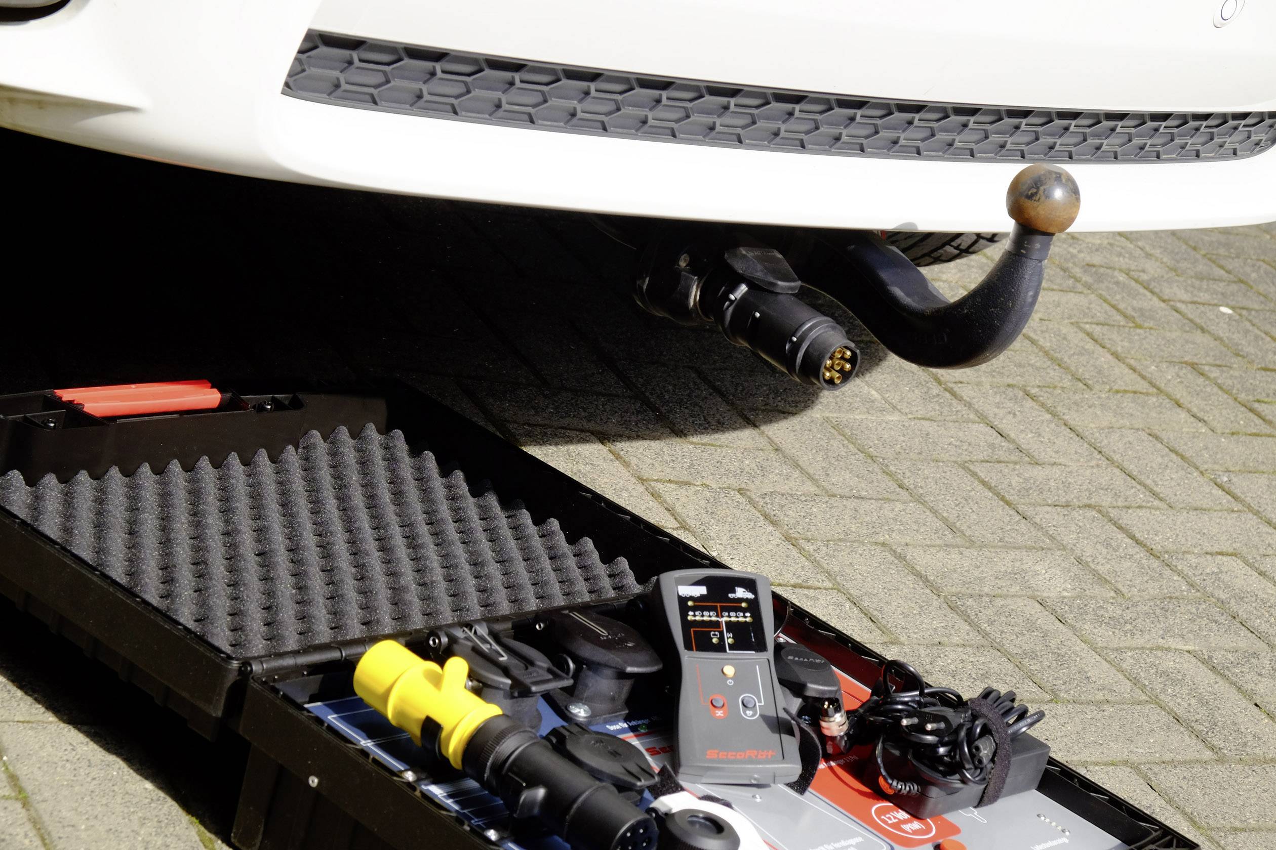 A white car with a tow bar is parked on cobblestones. In the foreground, an open toolbox containing various car accessories is visible.