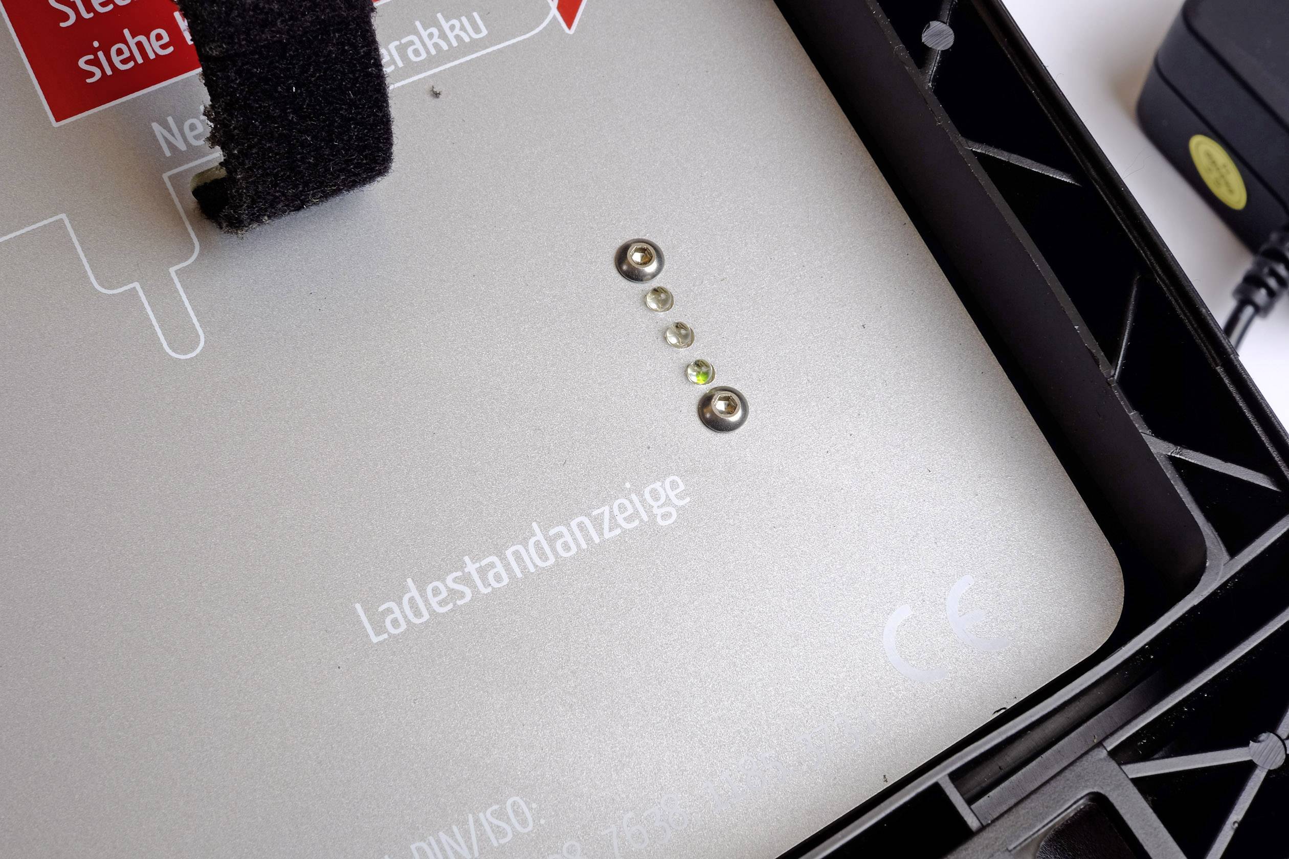 A silver device with four illuminated LEDs, the word 'charge level indicator' is visible. Part of a plug can be seen in the background.