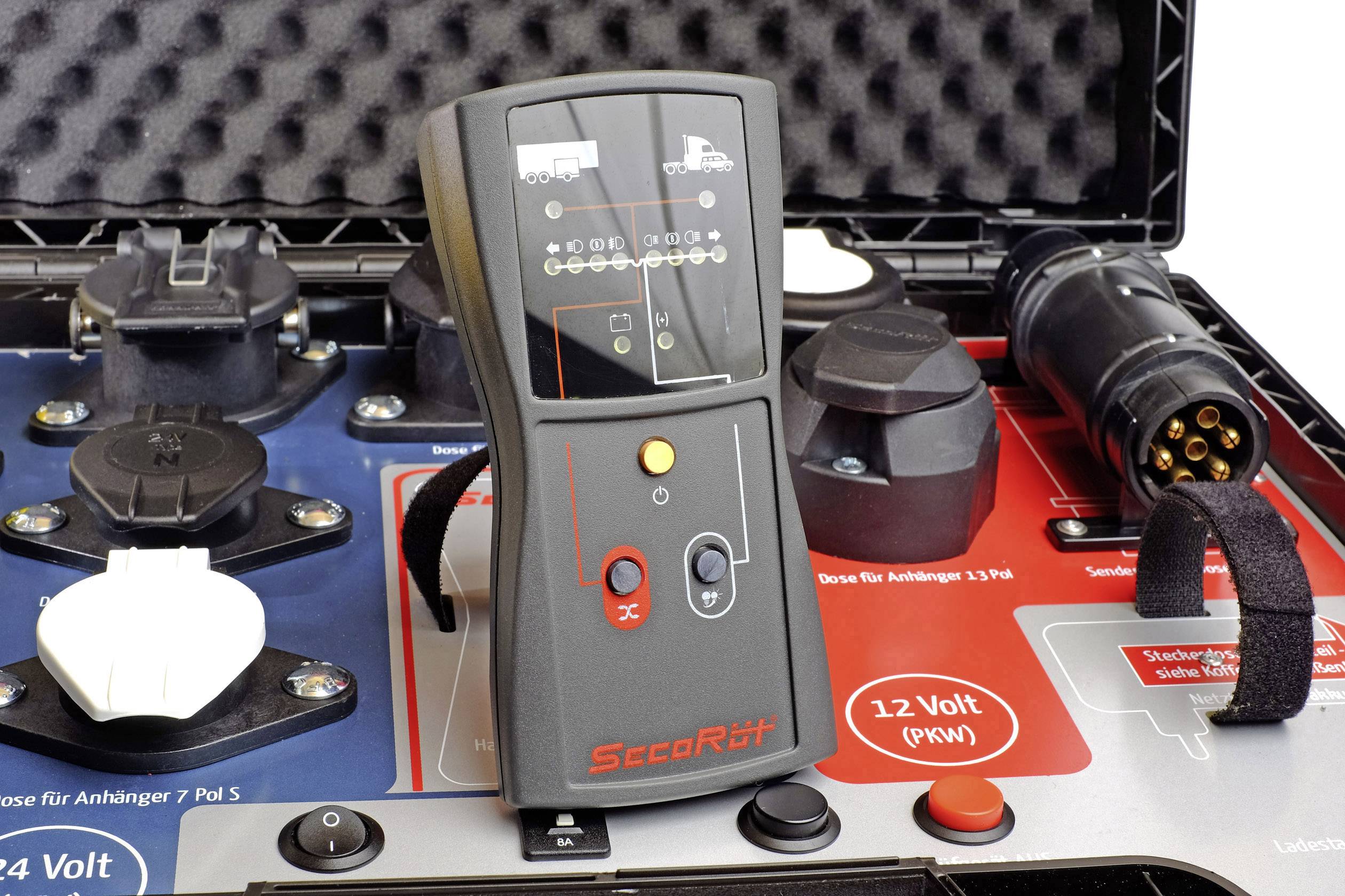 A black test device with multiple LED indicators and buttons lies in a case, surrounded by various connection plugs.