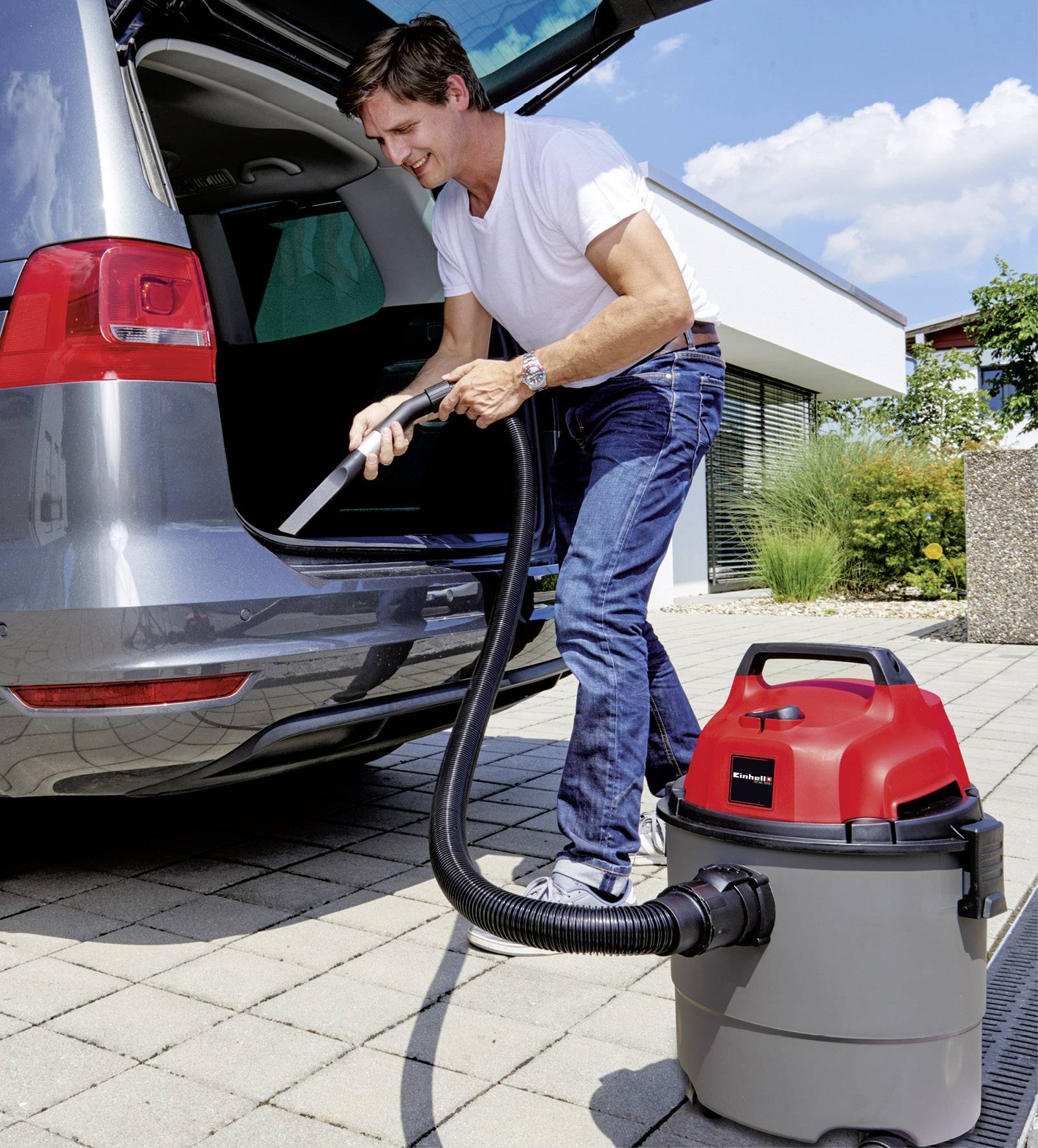 A man is vacuuming the interior of a car with an industrial vacuum cleaner on a paved driveway in front of a modern house.